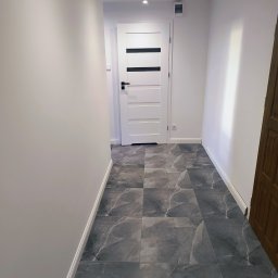 Sergi_job - Wnętrze korytarza z szarymi płytkami imitującymi marmur na podłodze. Białe ściany i drzwi z czarnymi akcentami. Oświetlenie sufitowe punktowe.