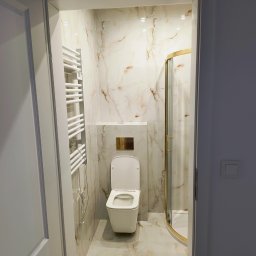 Sergi_job - Wykończona łazienka z kabiną prysznicową ze złotymi detalami, toaletą i grzejnikiem drabinkowym, widoczna zza otwartych drzwi. Marmurowe płytki na ścianach i podłodze.