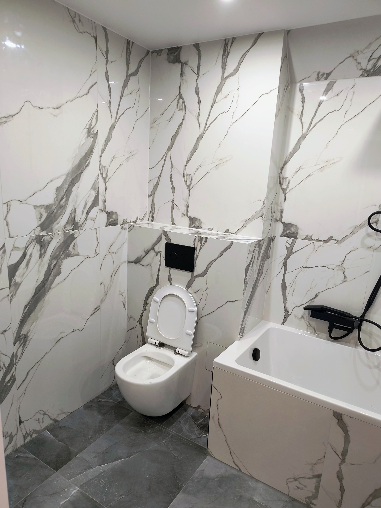 Wykończona łazienka w bieli i szarości: marmurowe płytki ścienne, szara podłoga, biała wanna i toaleta z czarnym panelem. Nowoczesny design.
