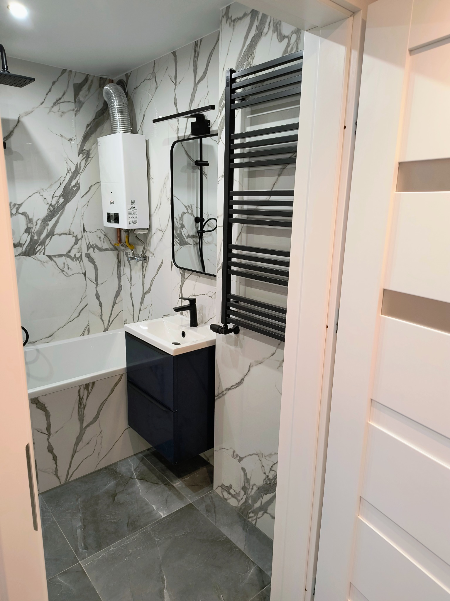 Wykończona łazienka z marmurowymi płytkami, zabudowaną umywalką w kolorze granatowym i czarnym grzejnikiem drabinkowym. Widoczny bojler i białe drzwi.