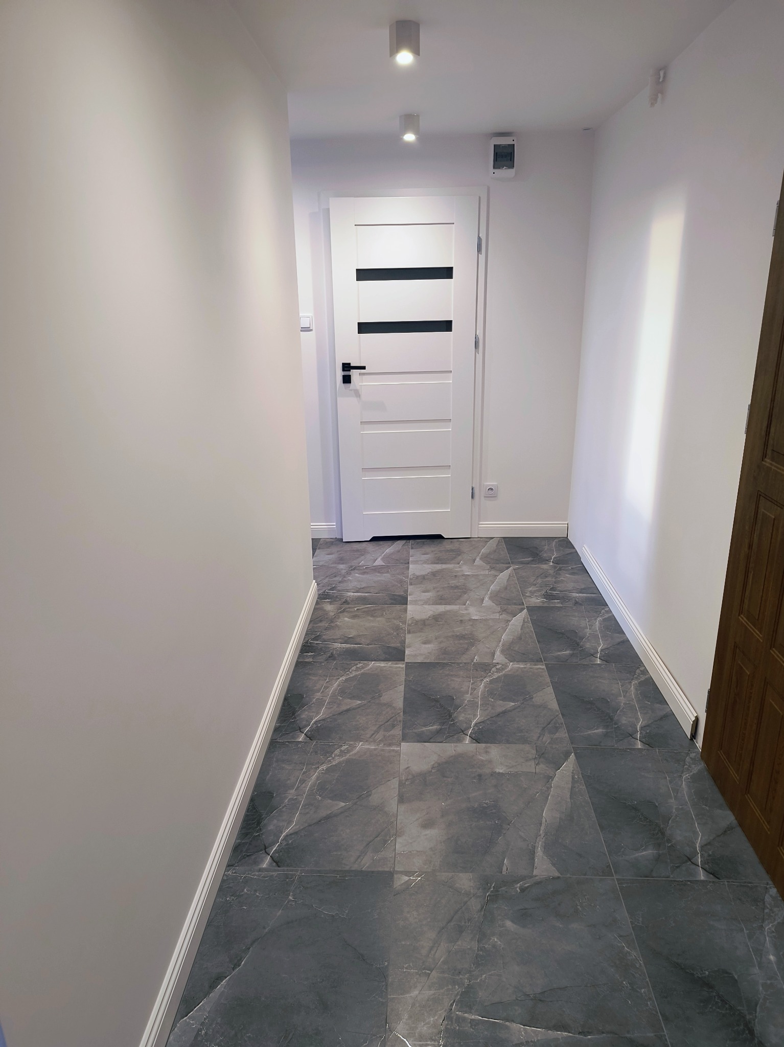 Wnętrze korytarza z szarymi płytkami imitującymi marmur na podłodze. Białe ściany i drzwi z czarnymi akcentami. Oświetlenie sufitowe punktowe.