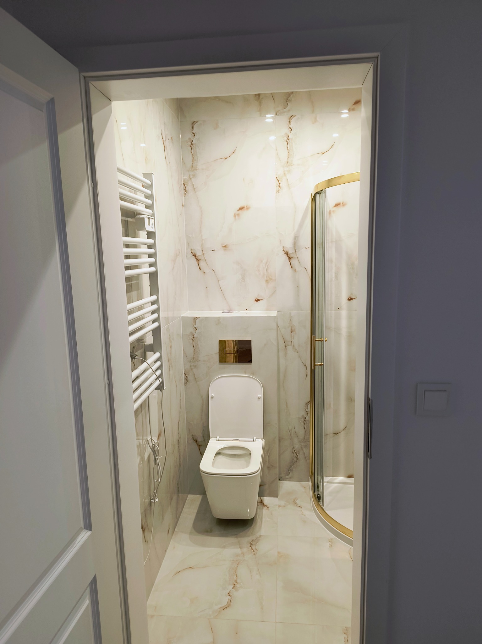 Wykończona łazienka z kabiną prysznicową ze złotymi detalami, toaletą i grzejnikiem drabinkowym, widoczna zza otwartych drzwi. Marmurowe płytki na ścianach i podłodze.