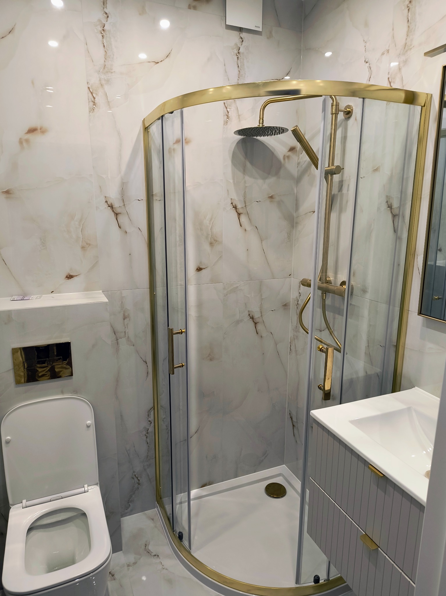 Wykończona łazienka z kabiną prysznicową w złotej ramie, umywalką z szarą szafką i toaletą. Płytki imitujące marmur na ścianach i podłodze.