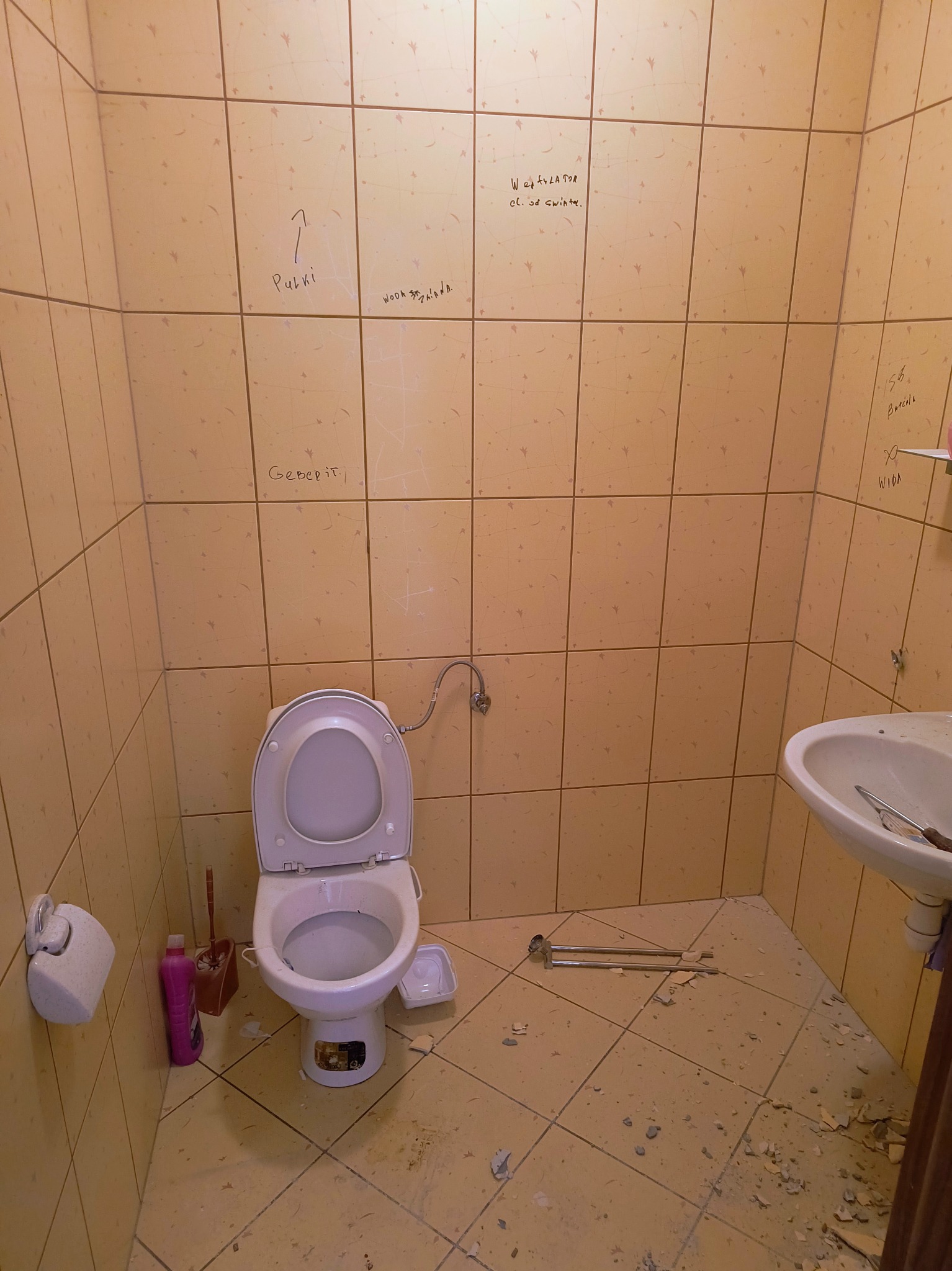 Łazienka w trakcie demontażu płytek, z widoczną toaletą, umywalką i pozostałościami po pracach. Na ścianach widoczne napisy, a na podłodze gruz.