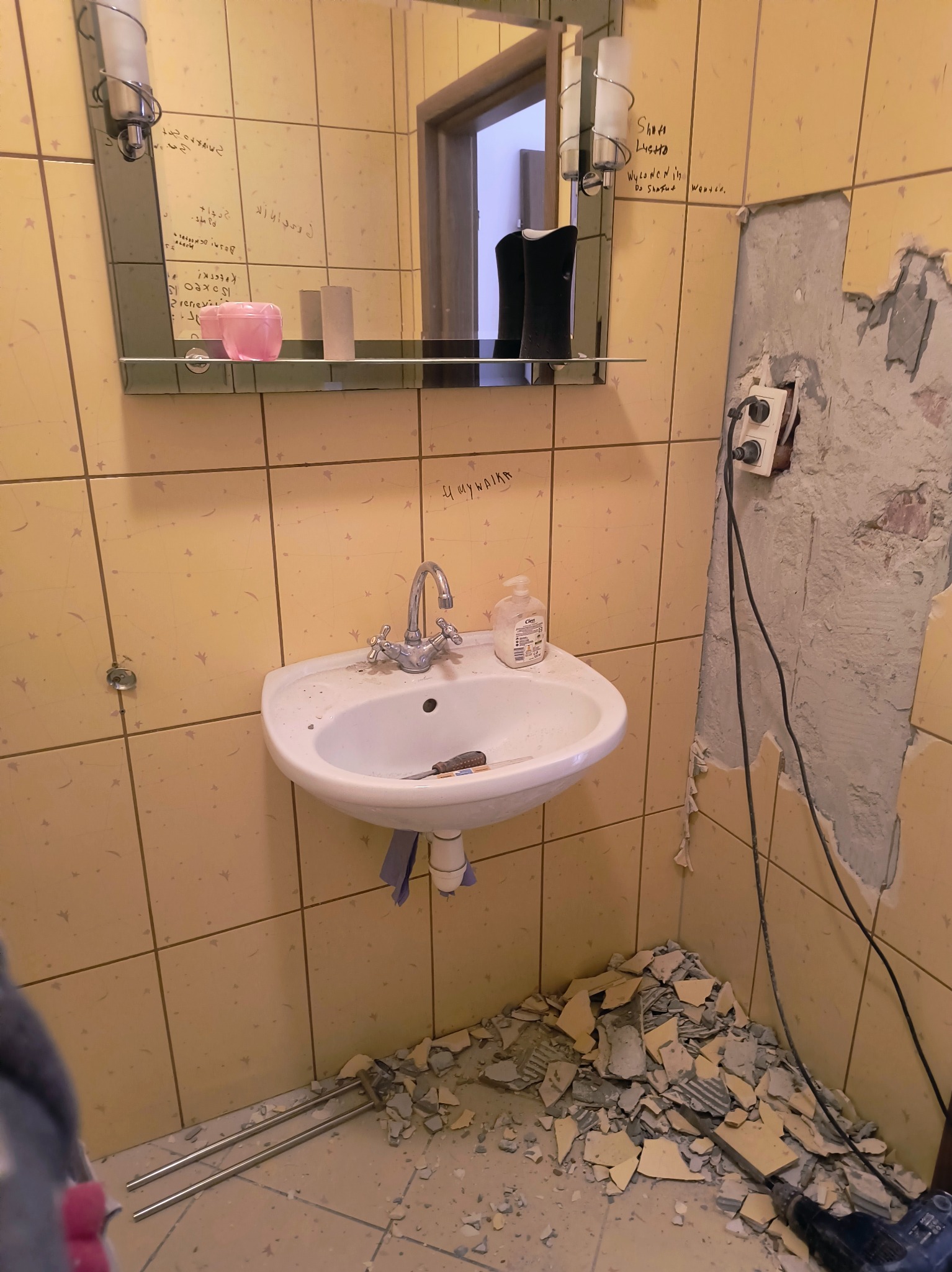 Żółta łazienka w trakcie demontażu płytek, widoczne narzędzia i gruz. Lustro z napisami, umywalka z kranem. Gniazdko elektryczne odsłonięte, ściany częściowo bez płytek.