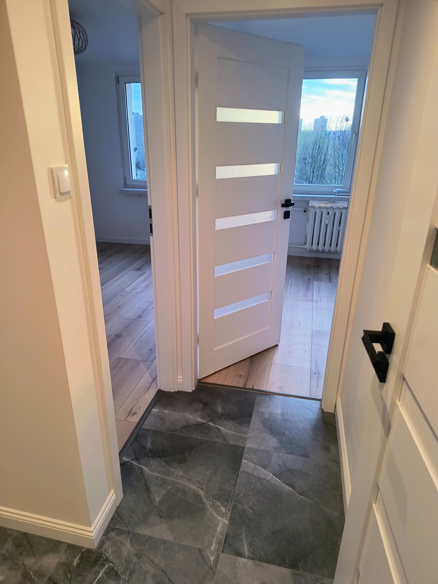 Nowoczesne, białe drzwi z przeszkleniami, czarne klamki. Szara, marmurkowa podłoga w przedpokoju, widok na parkiet w pokoju. Jasne, minimalistyczne wnętrze.