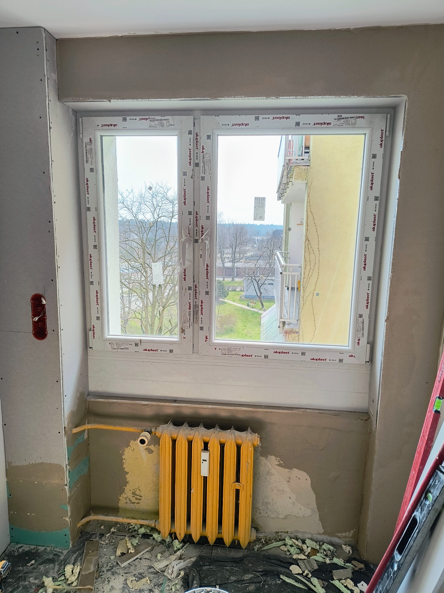 Wnęka okienna w trakcie prac wykończeniowych: zamontowane okno PCV, obudowa z płyt gipsowo-kartonowych, żółty grzejnik żeberkowy. Widok na zewnątrz.