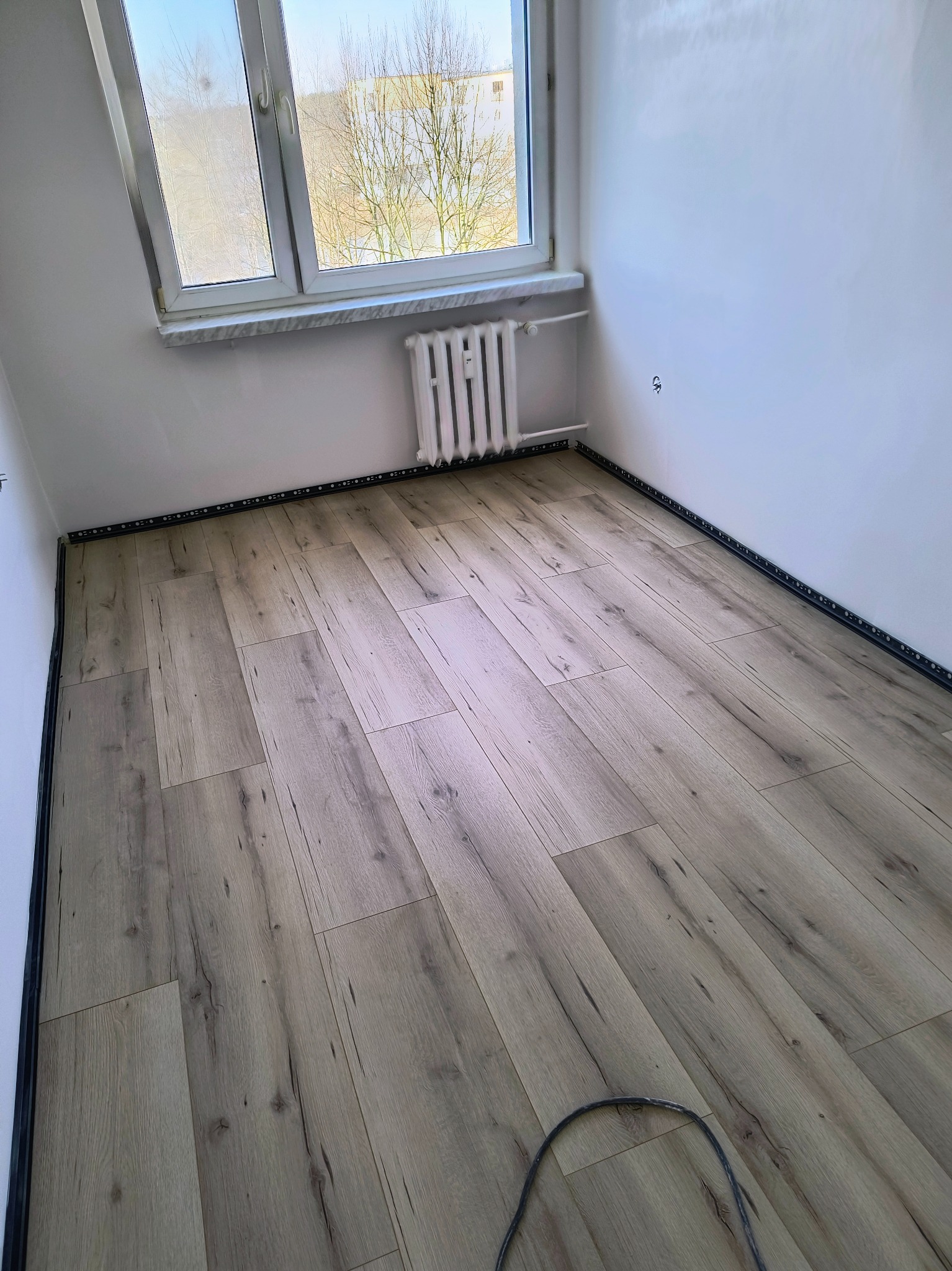 Jasne panele podłogowe ułożone w małym pokoju z grzejnikiem pod oknem. Listwy przypodłogowe zamontowane. Widoczny kabel na podłodze w rogu.