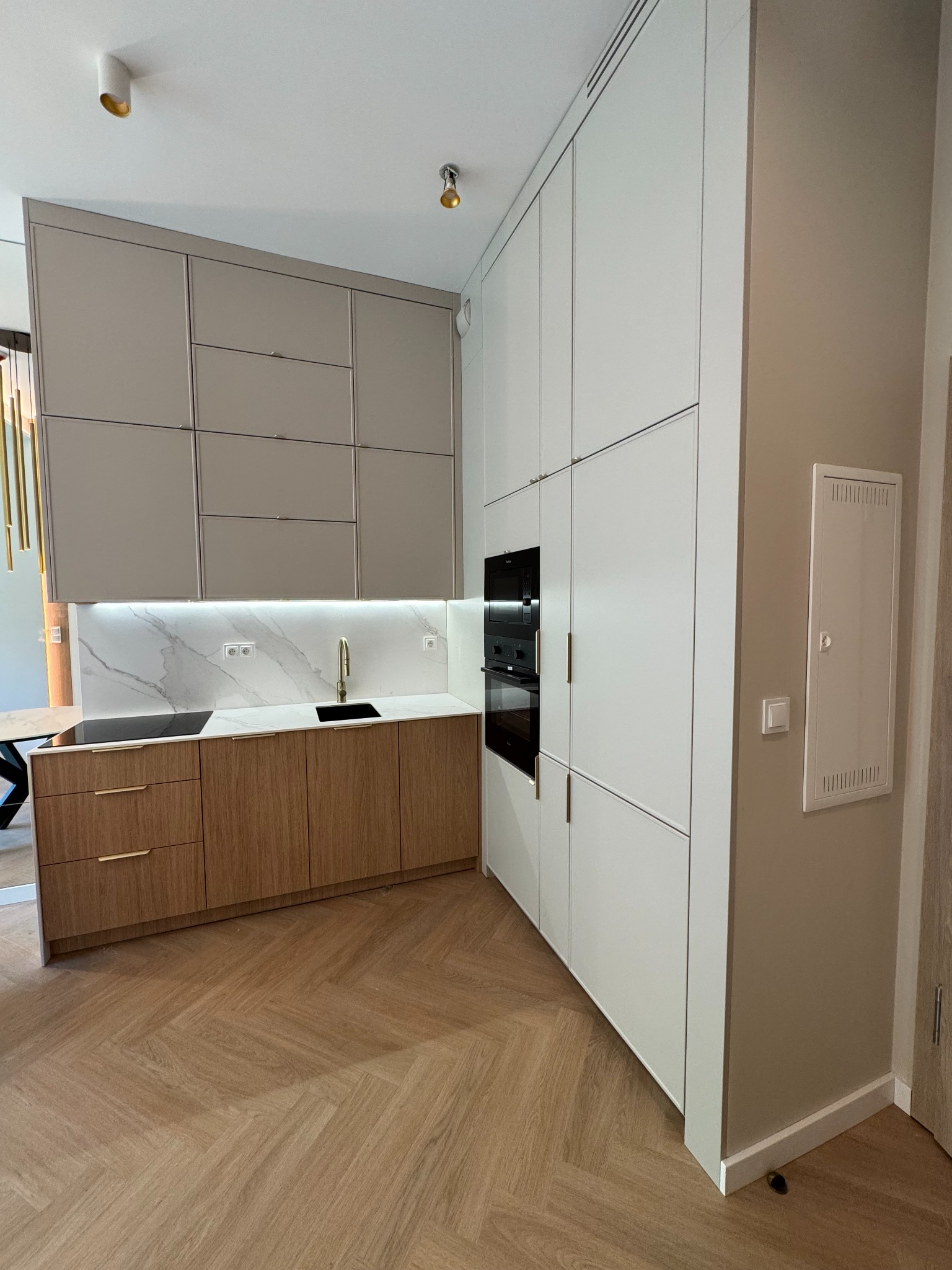 Nowoczesna kuchnia z drewnianymi frontami szafek dolnych i białymi górnymi, podświetlany blat z marmurowym wzorem, parkiet w jodełkę, piekarnik w zabudowie.