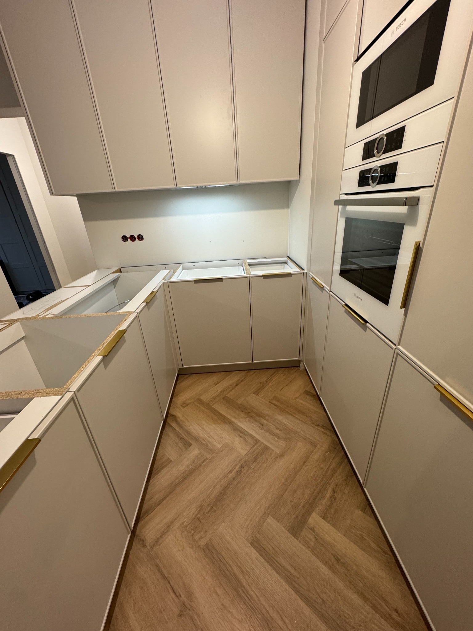 Montaż jasnych szafek kuchennych z złotymi uchwytami, w tle zabudowa piekarnika marki Bosch. Ułożona podłoga z paneli w jodełkę. Widać niedokończony montaż.