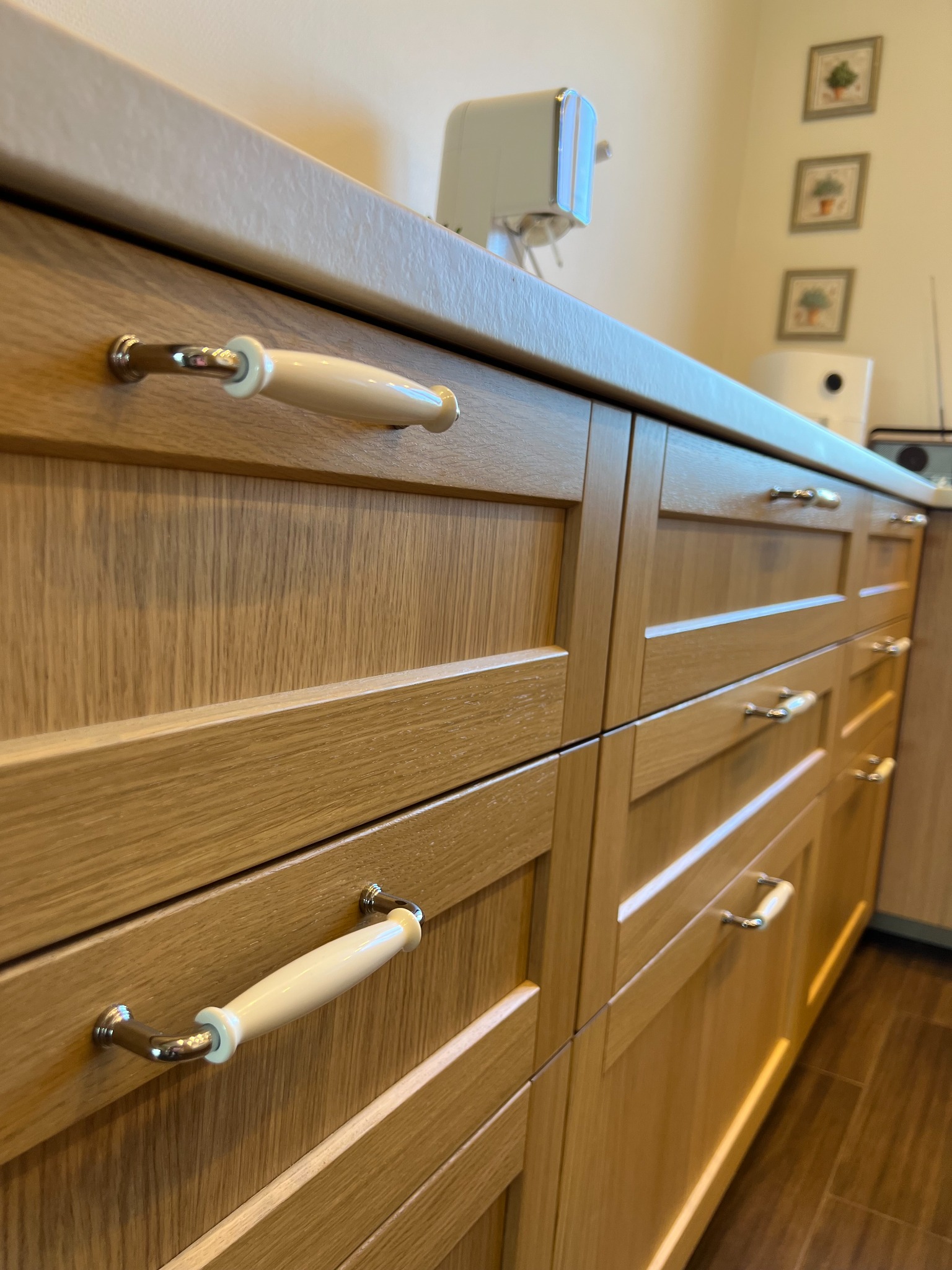 Fronty szuflad z jasnego drewna z kremowymi uchwytami w kuchni z beżowym blatem. W tle ekspres do kawy i dekoracje ścienne.