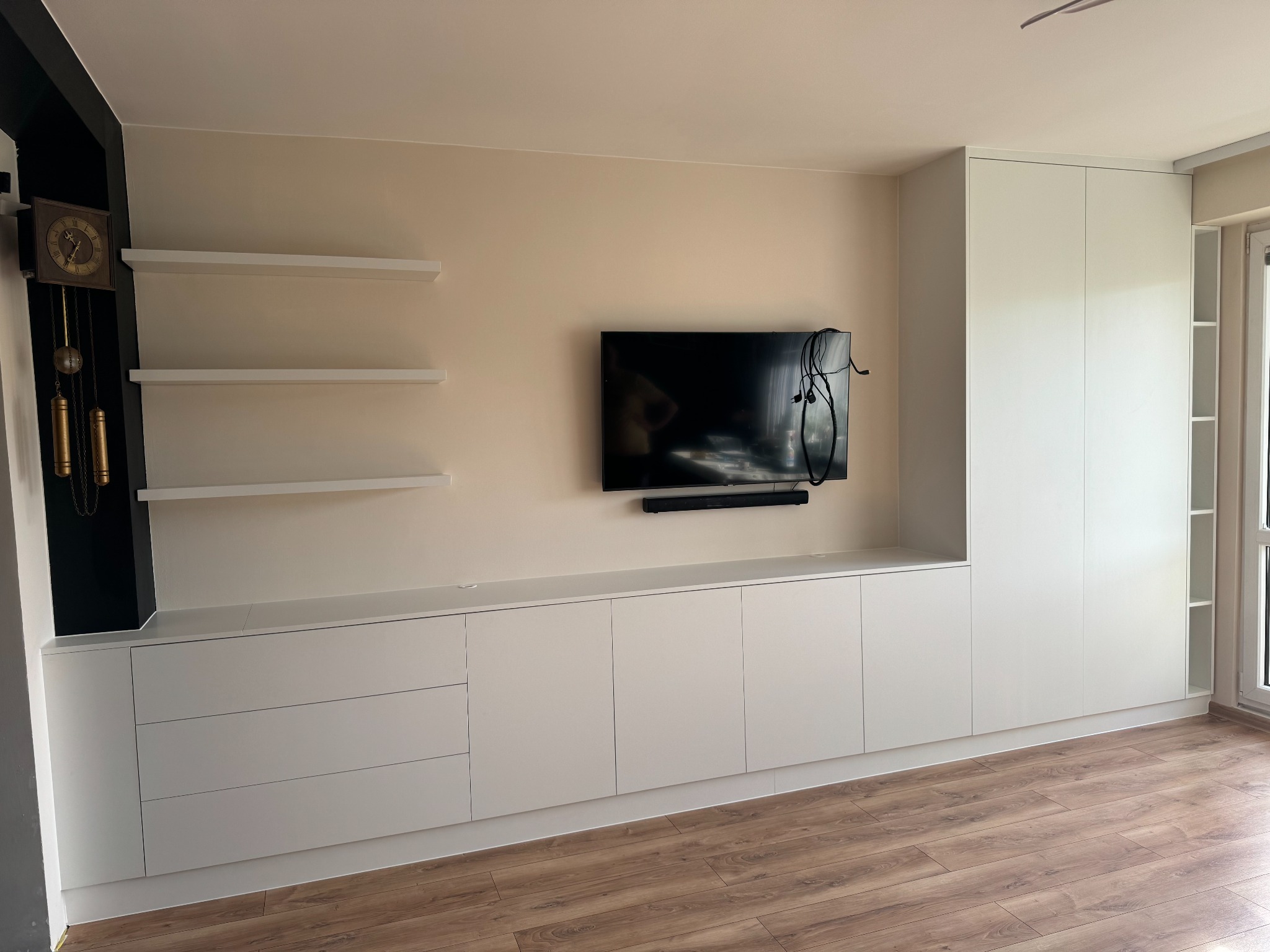 Biała zabudowa meblowa z szafkami i półkami, z telewizorem na ścianie. Widoczny drewniany zegar i panele podłogowe. Minimalistyczny design, jasne wnętrze.