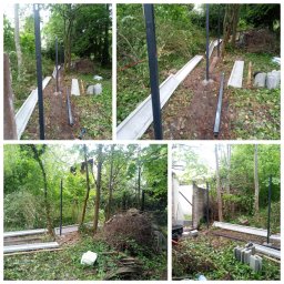 Drzewo PL 76 - Montaż ogrodzenia panelowego w zarośniętym ogrodzie: czarne słupki, betonowe podmurówki, narzędzia w tle. Etap budowy, widok na zieleń i elementy konstrukcyjne.