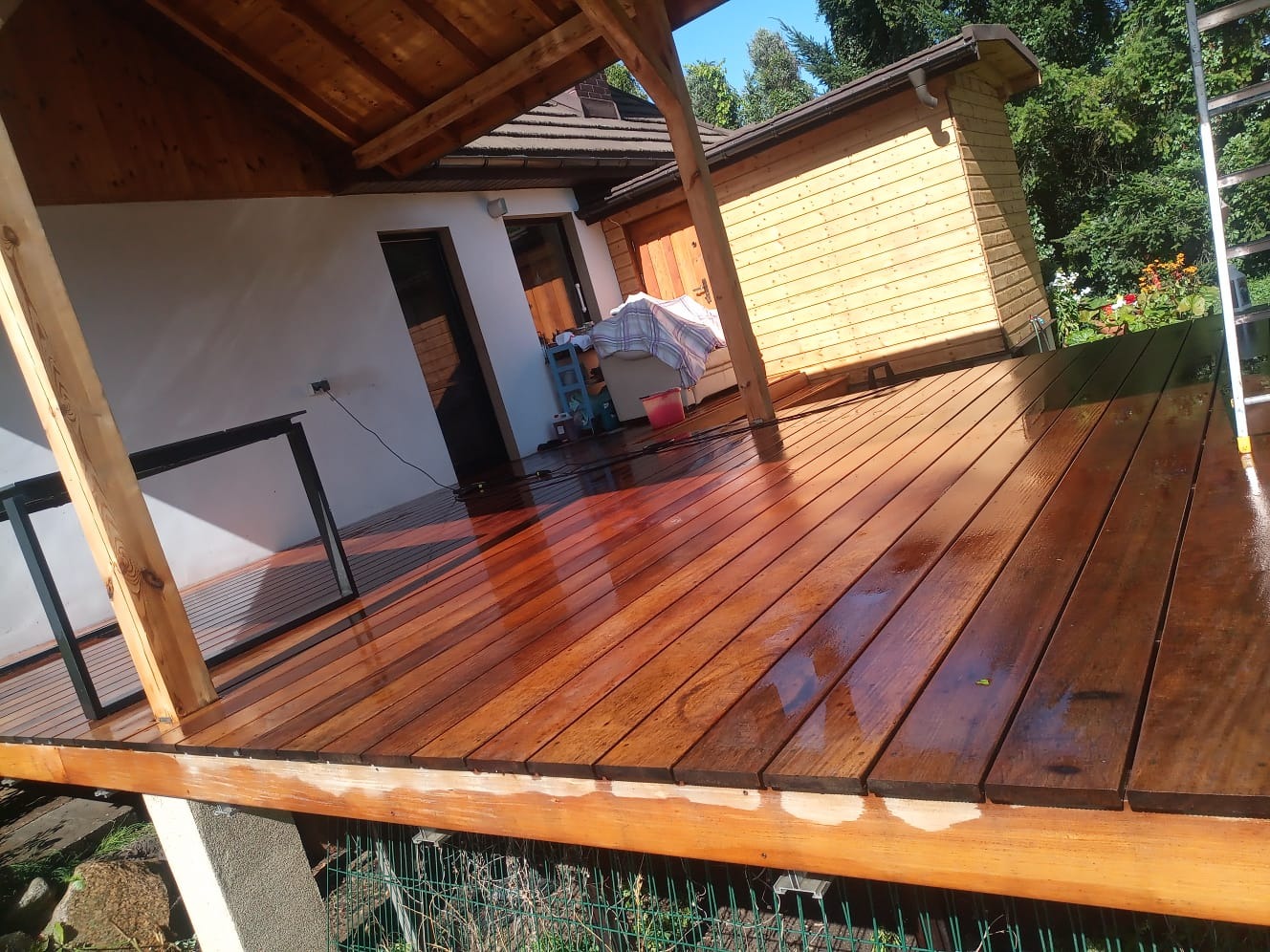 Świeżo polakierowany, drewniany taras przydomowy z widoczną konstrukcją zadaszenia, fragmentem elewacji i drabiny. Mokra powierzchnia odbija światło.