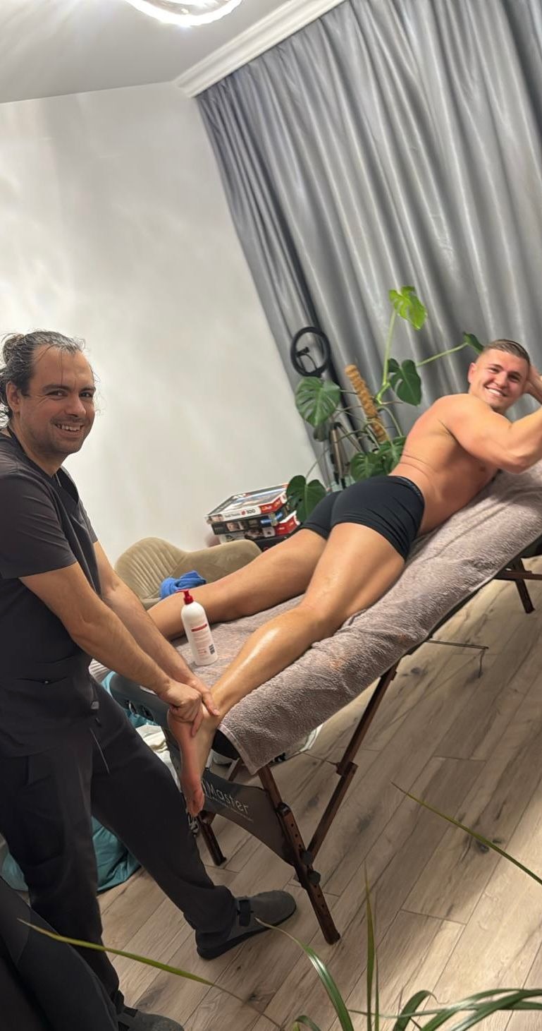 Mężczyzna w trakcie masażu stóp przez masażystę na leżance w gabinecie. Pacjent uśmiechnięty, w tle roślina doniczkowa i zasłony.