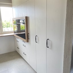 PPHU Goralczyk - Biała zabudowa kuchenna z czarnymi uchwytami i wbudowanym piekarnikiem w drewnianej ramie, widok z boku, jasne płytki podłogowe, okno z widokiem na zieleń.