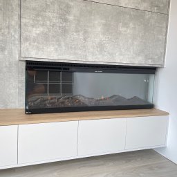 PPHU Goralczyk - Nowoczesna zabudowa ściany z elektrycznym kominkiem i szafkami z białymi frontami. Betonowa okładzina ściany i drewniany blat dodają elegancji.