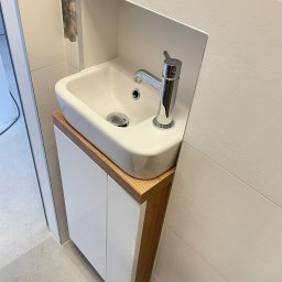 PPHU Goralczyk - Biała, nablatowa umywalka z chromowaną baterią, osadzona na szafce z drewnianym blatem w nowoczesnej łazience z płytkami imitującymi drewno. Minimalistyczny design.