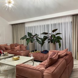 PPHU Goralczyk - Przestronny salon z narożną sofą w odcieniach różu, stolikami kawowymi i dużymi roślinami doniczkowymi przy oknie z zasłonami. Parkiet w jodełkę i nowoczesne oświetlenie.