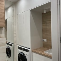 PPHU Goralczyk - Biała zabudowa meblowa z pralką i drewnianym blatem w łazience, widok z perspektywy. Projekt w Łomży z oświetleniem punktowym i lustrem powiększającym przestrzeń.