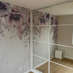 PPHU Goralczyk - Wnętrze pokoju z szafą przesuwną z lustrami, odbijającą białą szafkę nocną i okno. Na ścianie widoczna tapeta w kwiaty w odcieniach fioletu i różu.