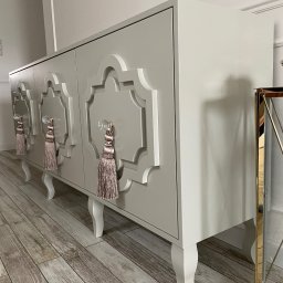 PPHU Goralczyk - Stylowa, beżowa komoda na nóżkach z ozdobnymi uchwytami i frezowanym wzorem na frontach, ustawiona na panelach podłogowych.