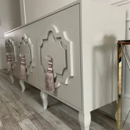 Stylowa, beżowa komoda na nóżkach z ozdobnymi uchwytami i frezowanym wzorem na frontach, ustawiona na panelach podłogowych.