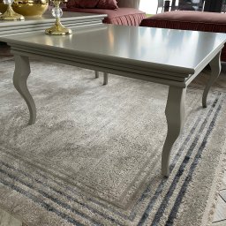 PPHU Goralczyk - Stylowy, szary stolik kawowy z dekoracyjnymi nogami na wzorzystym dywanie. W tle widoczny pluszowy puf i fragment podłogi z płytek imitujących drewno.