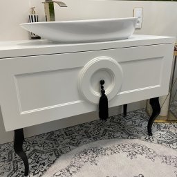 PPHU Goralczyk - Biała szafka łazienkowa z umywalką nablatową i dekoracyjnym uchwytem z frędzlami. Czarne, gięte nóżki, wzorzysta podłoga i akcesoria w tle.