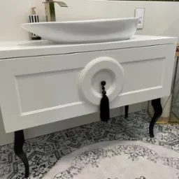 Biała szafka łazienkowa z umywalką nablatową i dekoracyjnym uchwytem z frędzlami. Czarne, gięte nóżki, wzorzysta podłoga i akcesoria w tle.