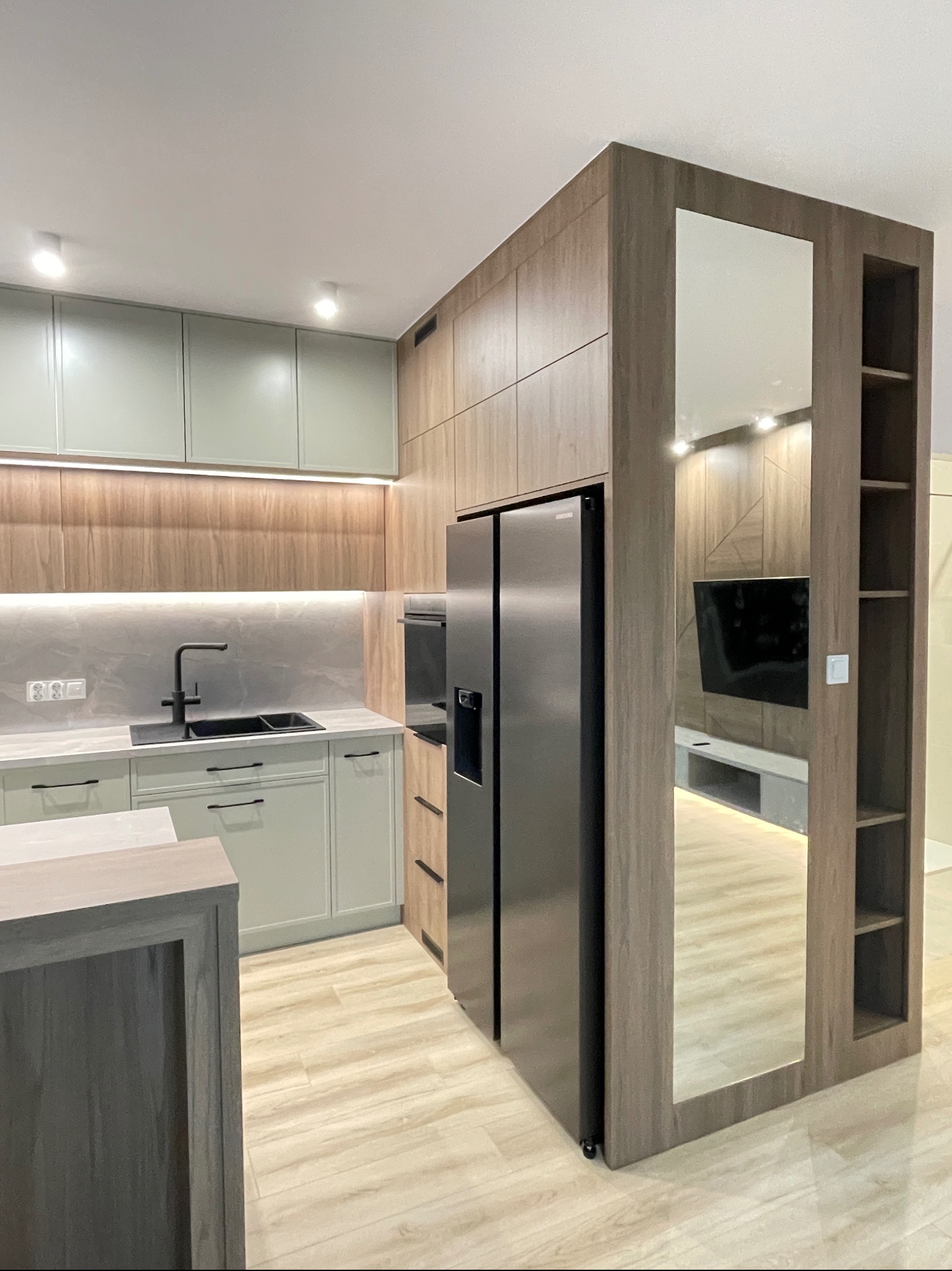 Nowoczesna kuchnia z drewnianymi frontami i szarymi szafkami, czarny zlew, lodówka side-by-side oraz lustro w zabudowie, odbijające wnętrze salonu.