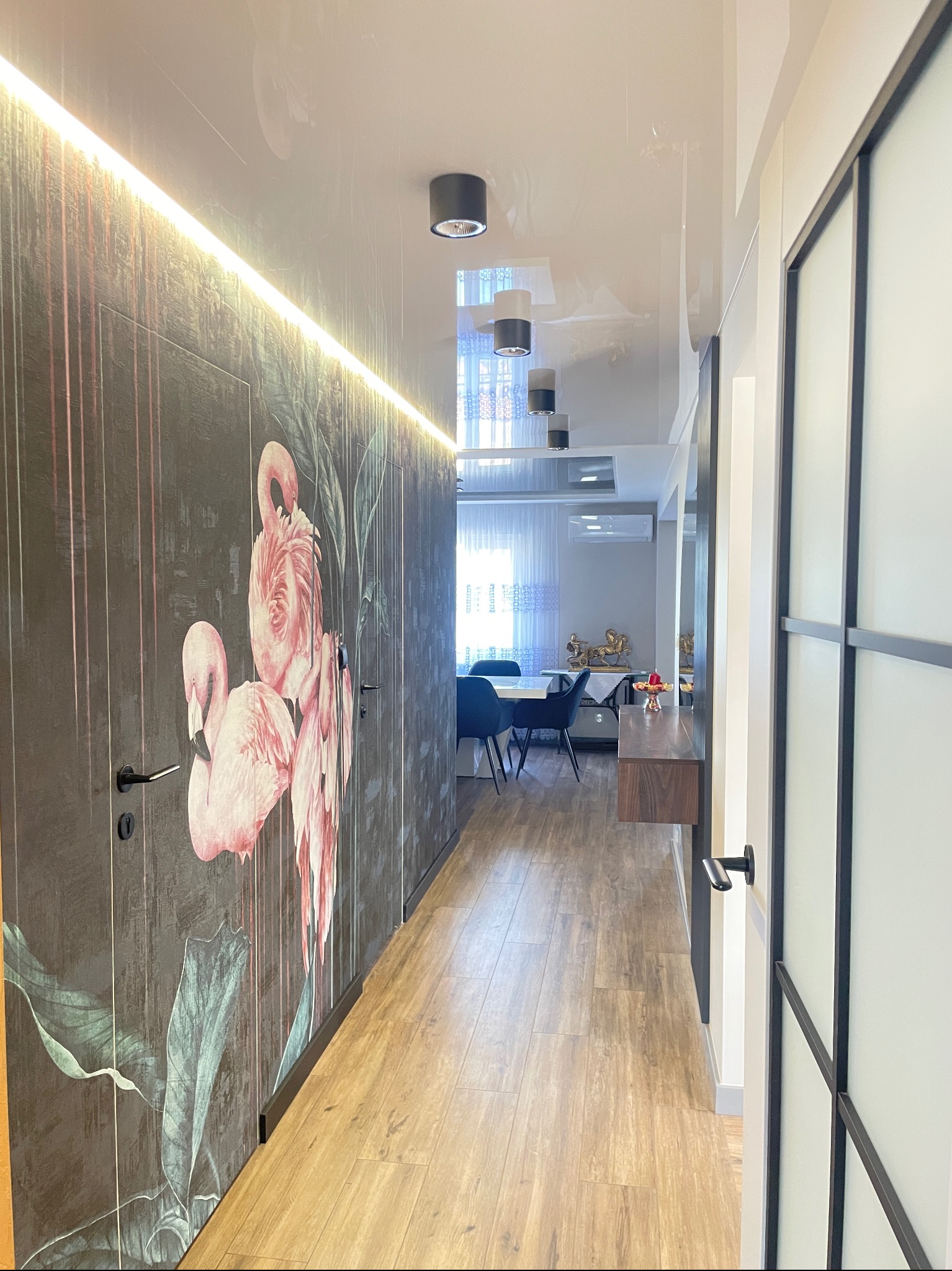 Nowoczesny przedpokój z szafą w zabudowie z motywem flamingów, oświetleniem LED i drewnianą podłogą. W tle widoczna jadalnia z niebieskimi krzesłami.