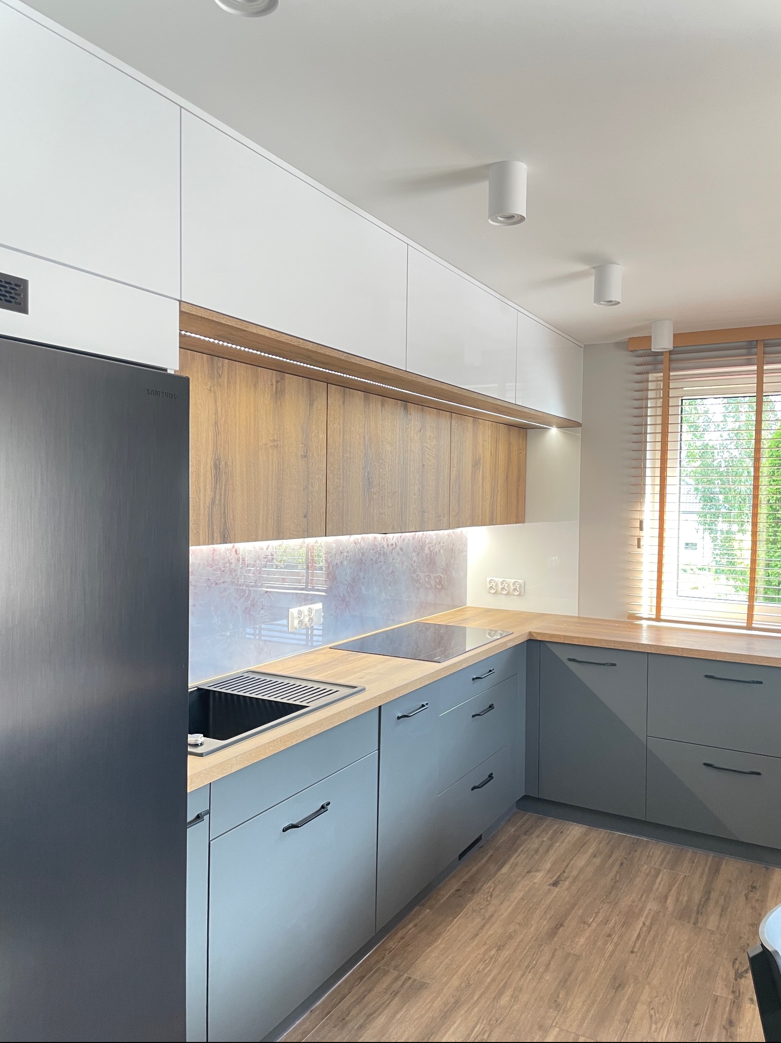 Nowoczesna kuchnia z szarymi szafkami, drewnianym blatem i białymi górnymi szafkami. Widoczne okno z drewnianymi żaluzjami i czarna lodówka Samsung.