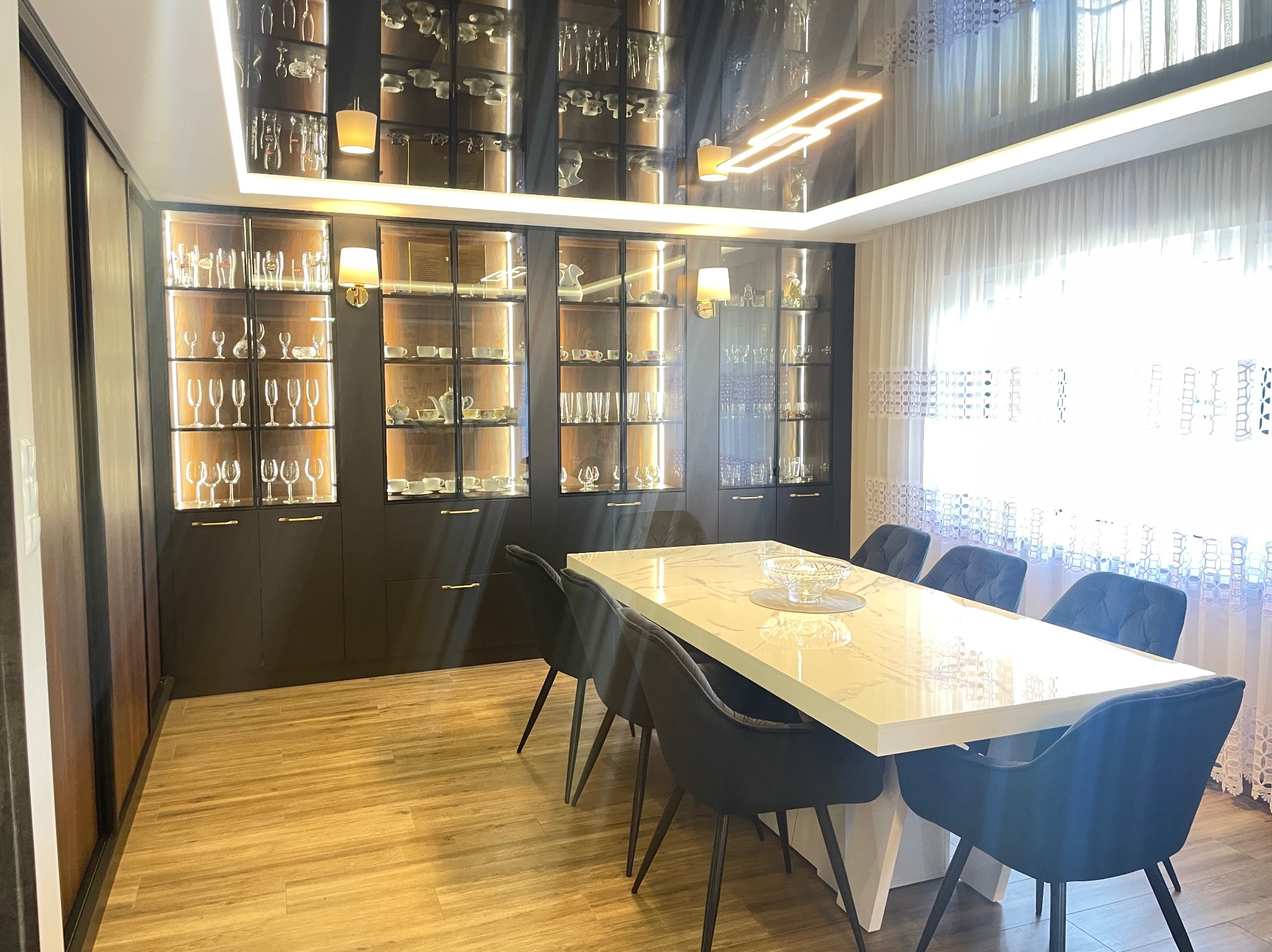 Elegancka jadalnia: biały stół z marmurowym blatem, welurowe krzesła, zabudowa z przeszklonymi szafkami z oświetleniem, parkiet. Nowoczesny design.