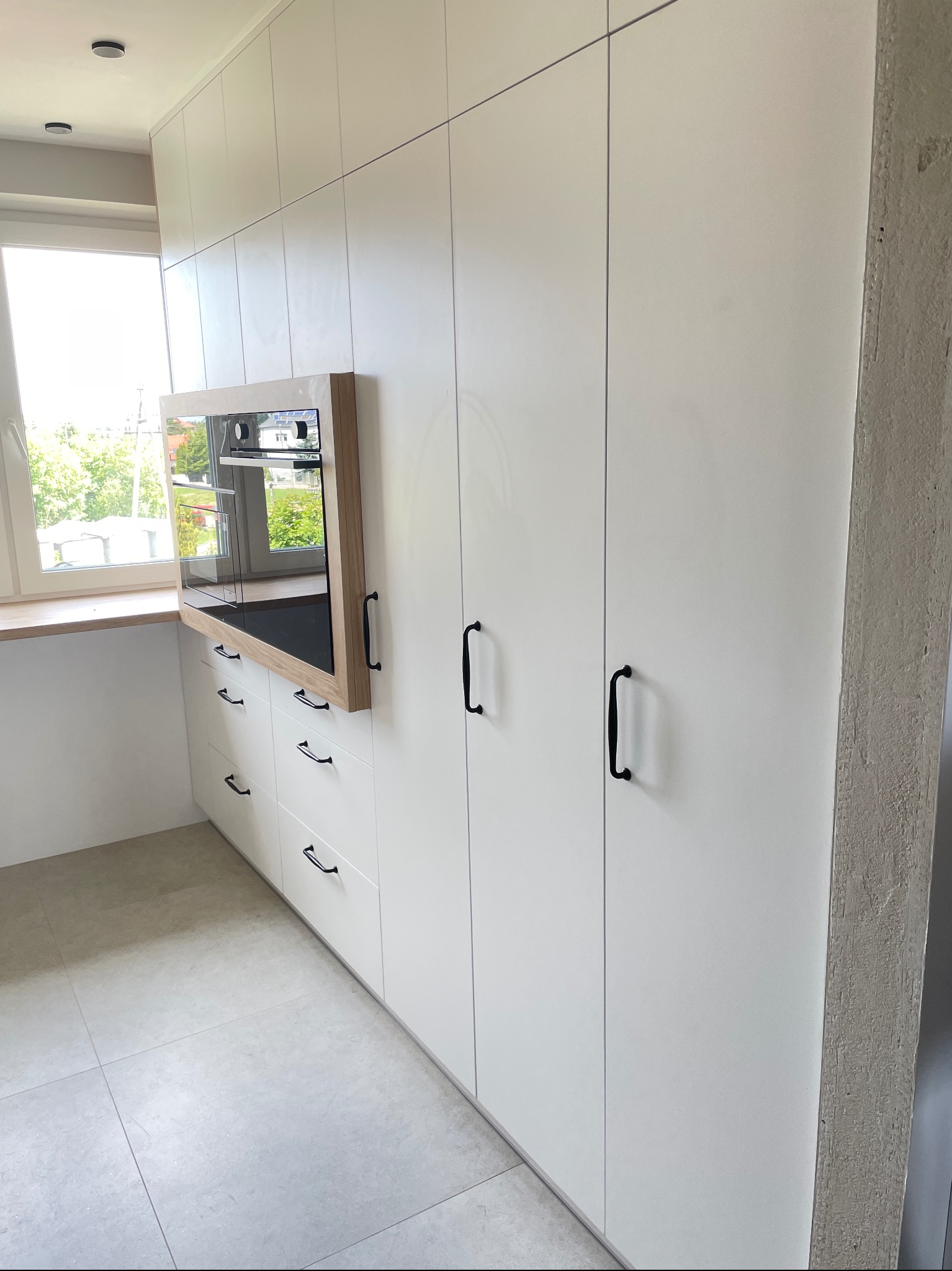 Biała zabudowa kuchenna z czarnymi uchwytami i wbudowanym piekarnikiem w drewnianej ramie, widok z boku, jasne płytki podłogowe, okno z widokiem na zieleń.