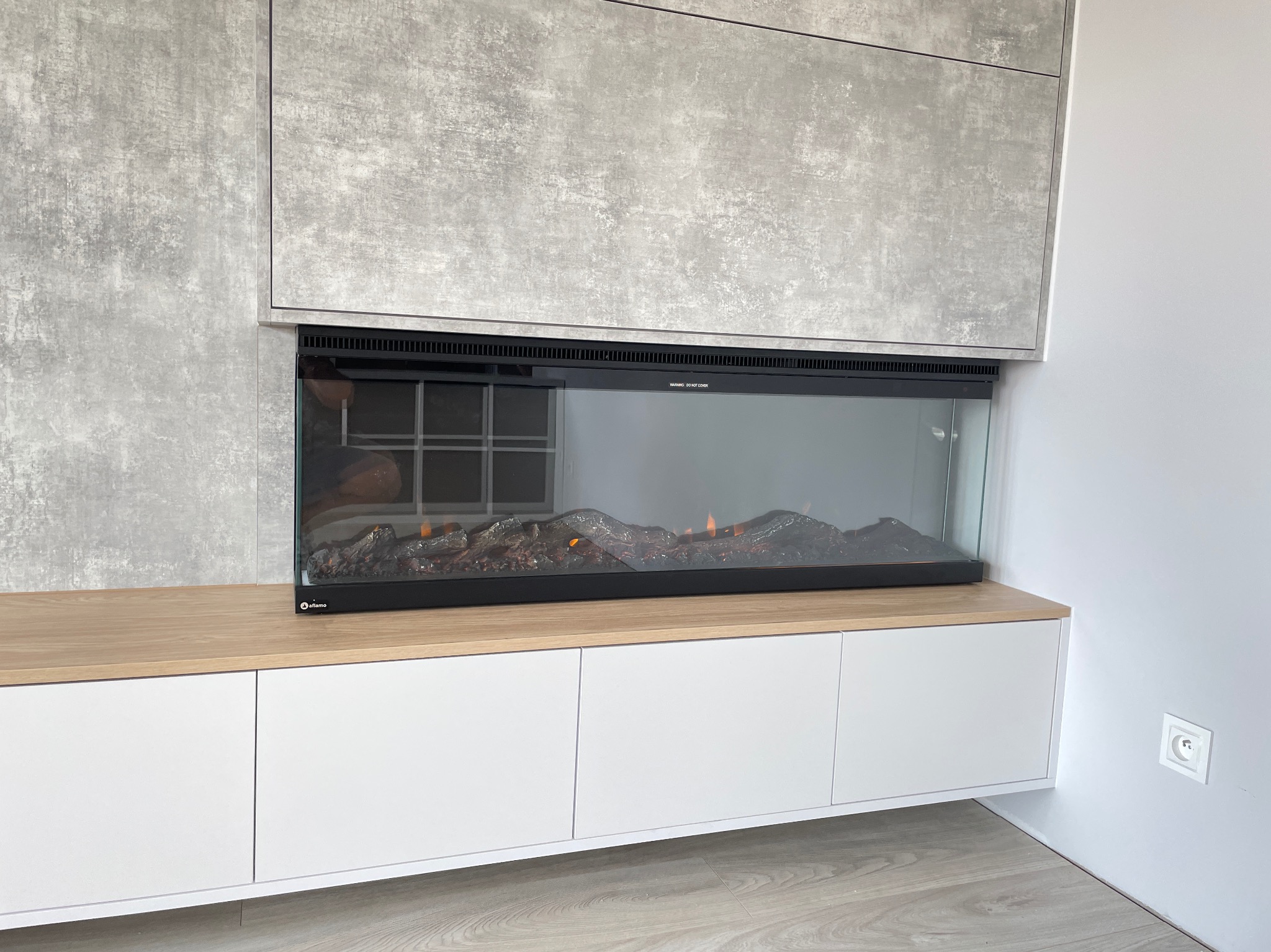 Nowoczesna zabudowa ściany z elektrycznym kominkiem i szafkami z białymi frontami. Betonowa okładzina ściany i drewniany blat dodają elegancji.