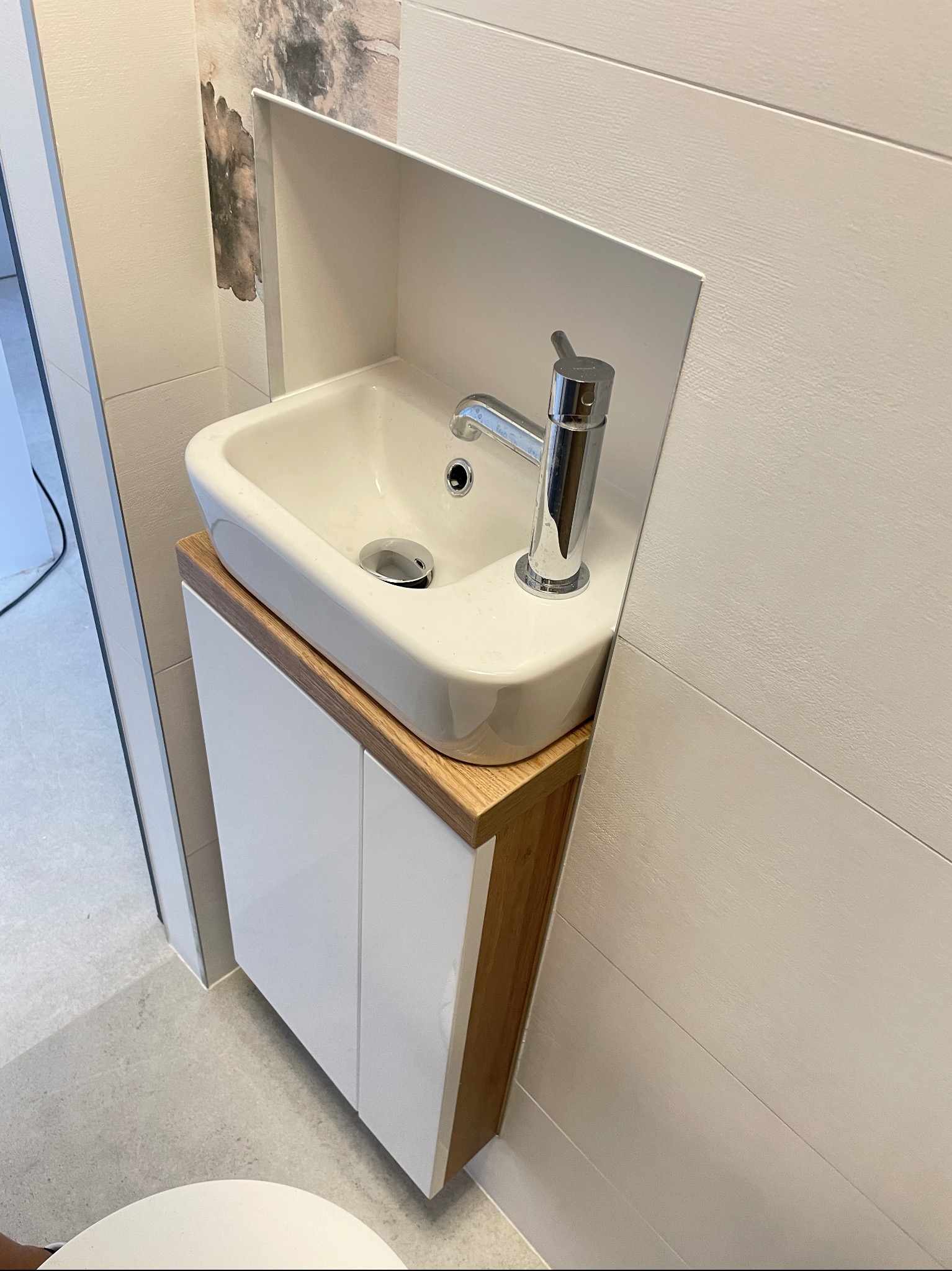 Biała, nablatowa umywalka z chromowaną baterią, osadzona na szafce z drewnianym blatem w nowoczesnej łazience z płytkami imitującymi drewno. Minimalistyczny design.