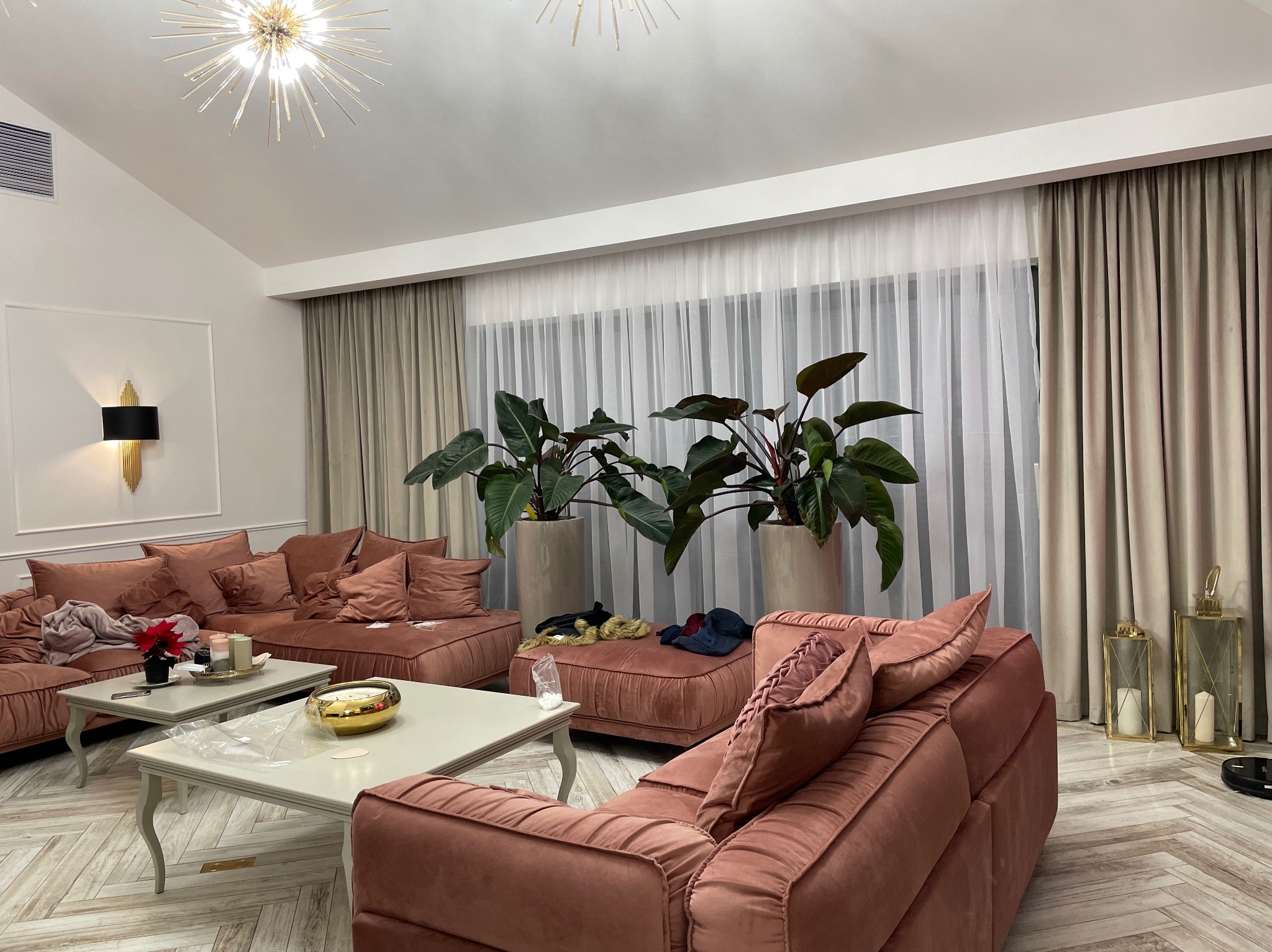 Przestronny salon z narożną sofą w odcieniach różu, stolikami kawowymi i dużymi roślinami doniczkowymi przy oknie z zasłonami. Parkiet w jodełkę i nowoczesne oświetlenie.