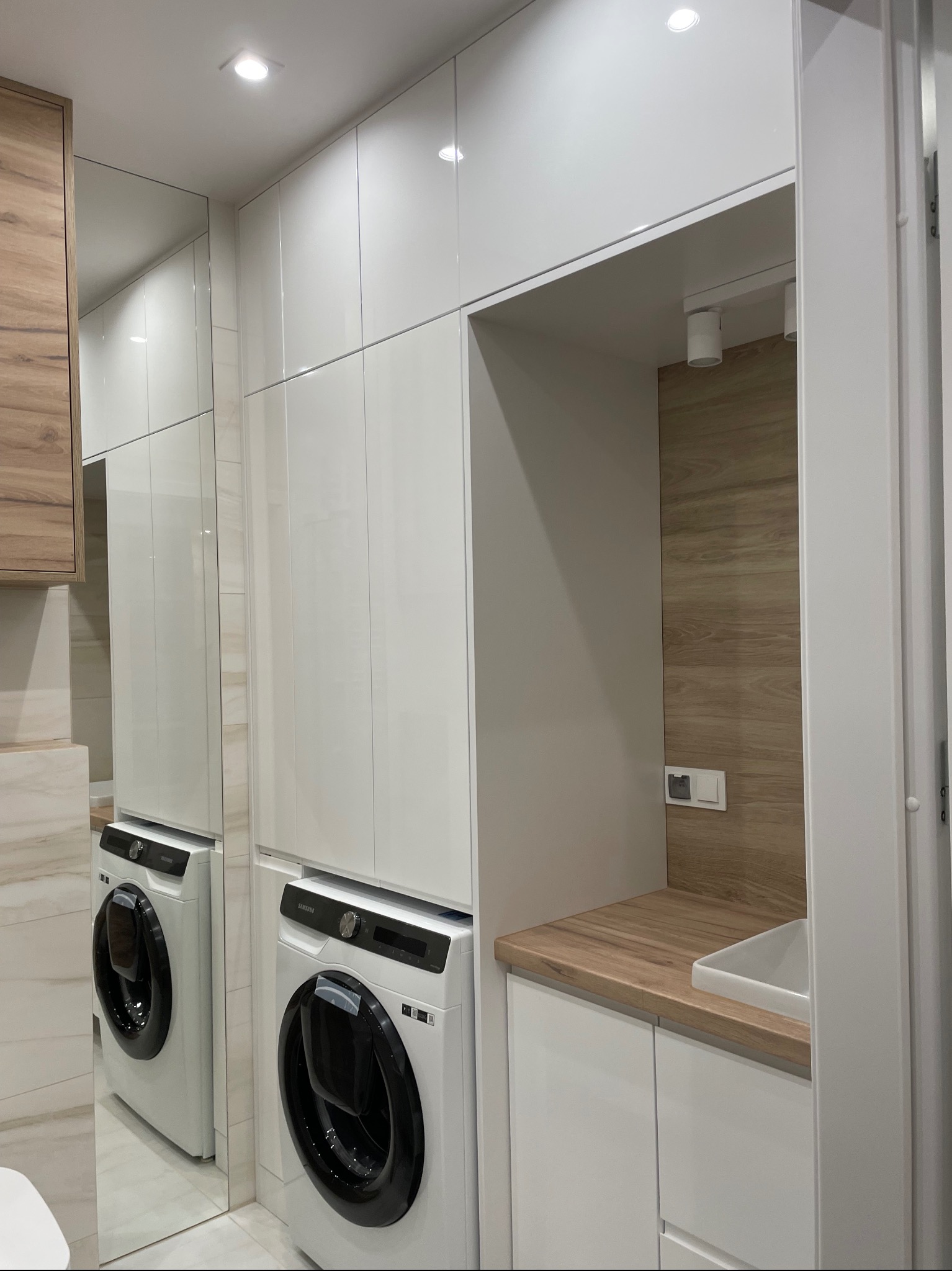 Biała zabudowa meblowa z pralką i drewnianym blatem w łazience, widok z perspektywy. Projekt w Łomży z oświetleniem punktowym i lustrem powiększającym przestrzeń.