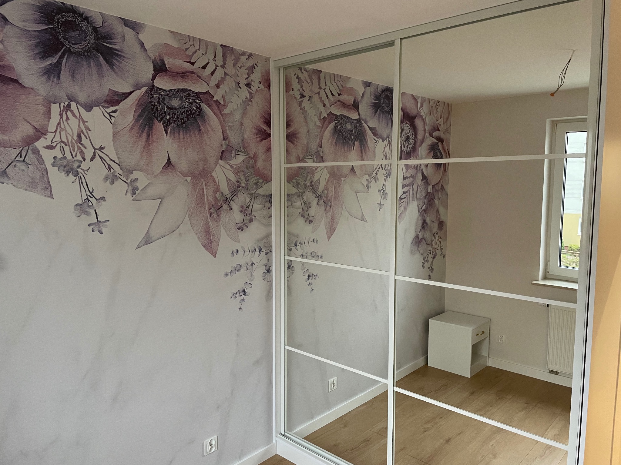 Wnętrze pokoju z szafą przesuwną z lustrami, odbijającą białą szafkę nocną i okno. Na ścianie widoczna tapeta w kwiaty w odcieniach fioletu i różu.