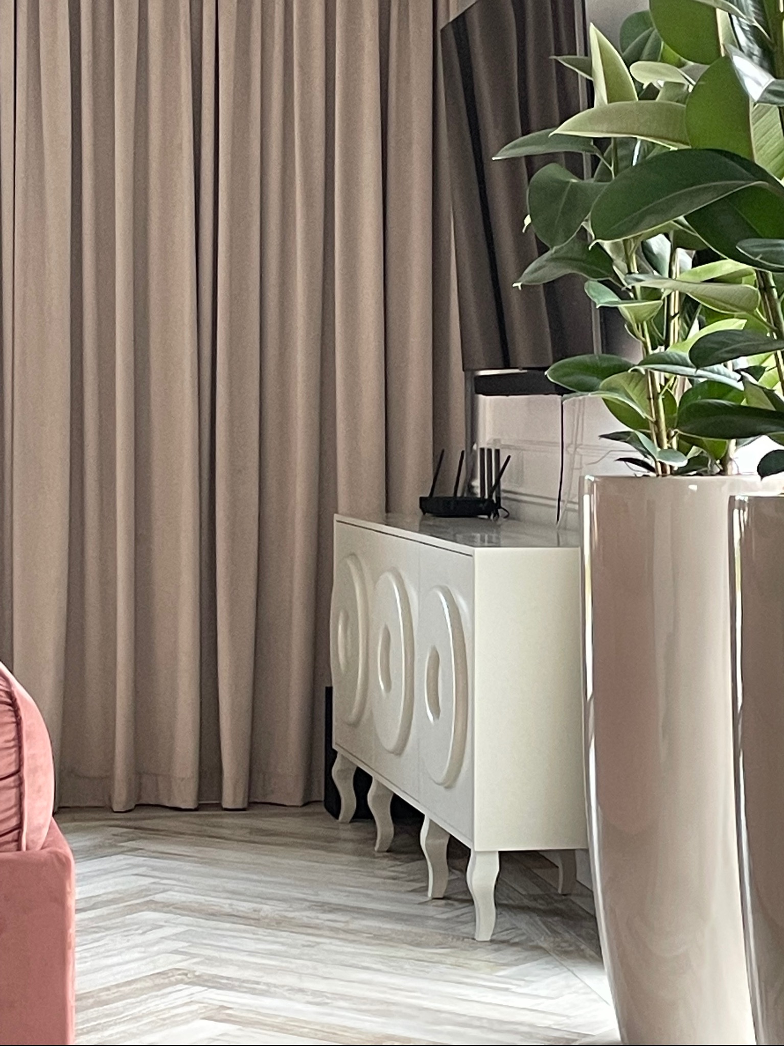 Elegancka komoda z dekoracyjnymi wycięciami na tle beżowych zasłon, uzupełniona donicą z dużą rośliną i widocznym routerem. Parkiet ułożony w jodełkę.