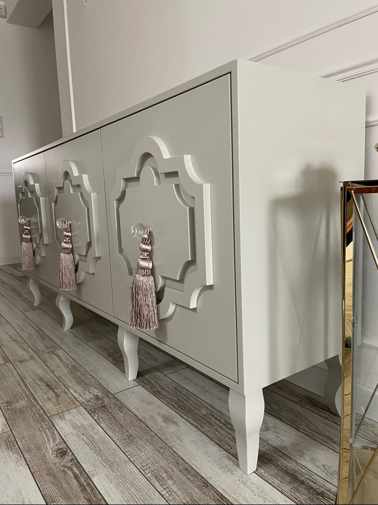 Stylowa, beżowa komoda na nóżkach z ozdobnymi uchwytami i frezowanym wzorem na frontach, ustawiona na panelach podłogowych.