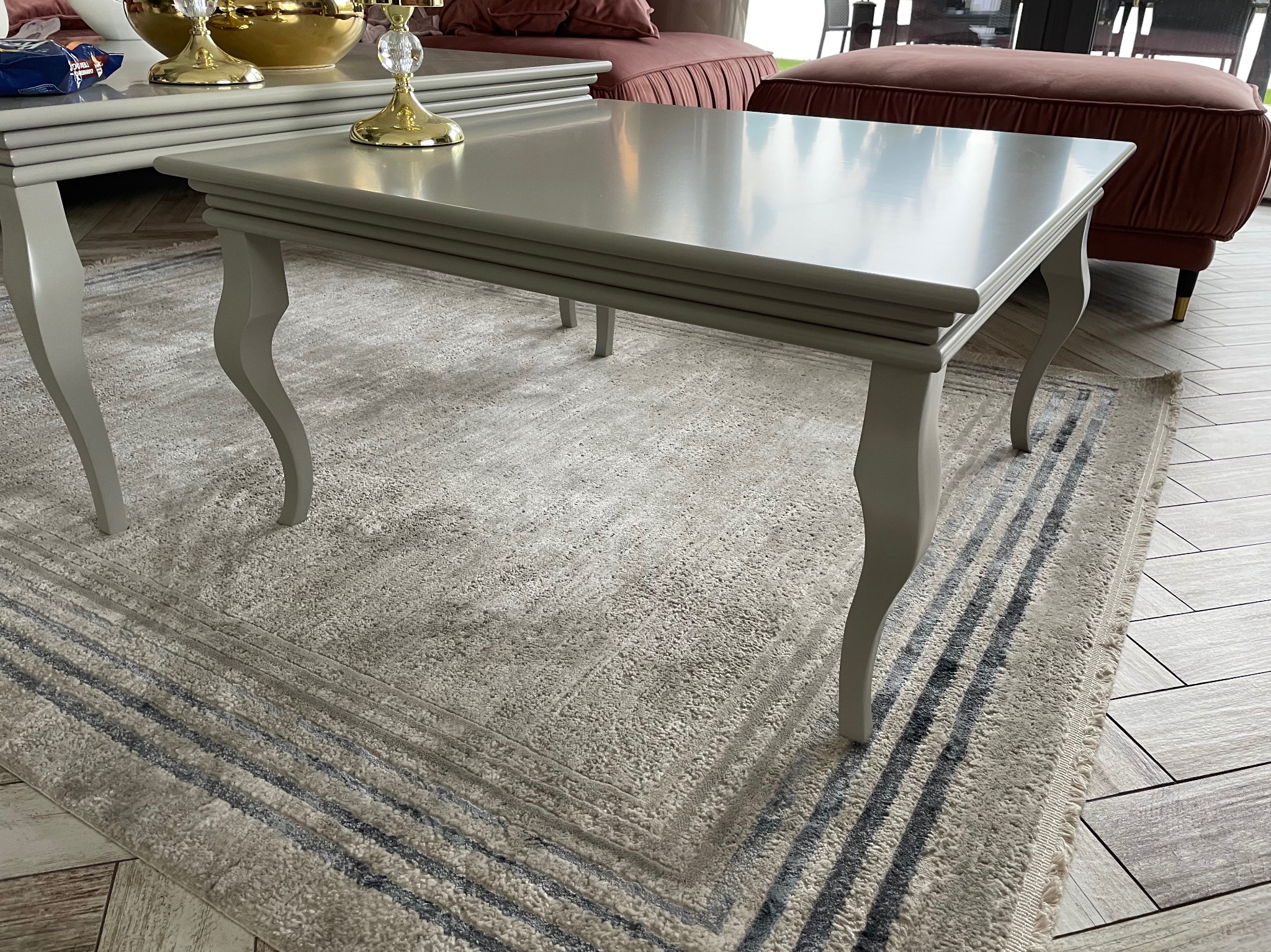 Stylowy, szary stolik kawowy z dekoracyjnymi nogami na wzorzystym dywanie. W tle widoczny pluszowy puf i fragment podłogi z płytek imitujących drewno.