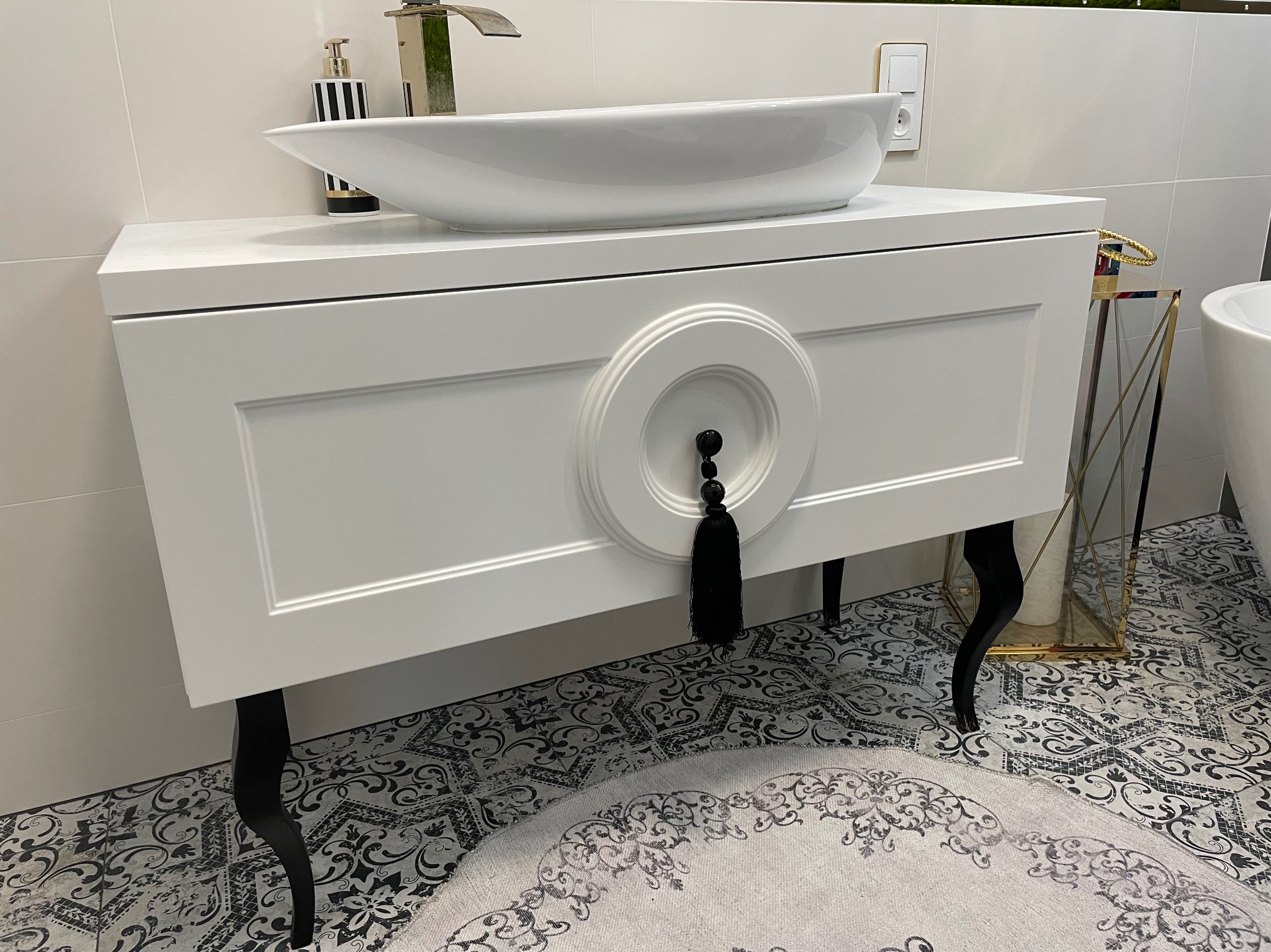 Biała szafka łazienkowa z umywalką nablatową i dekoracyjnym uchwytem z frędzlami. Czarne, gięte nóżki, wzorzysta podłoga i akcesoria w tle.