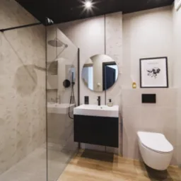 Beżowa łazienka z kabiną walk-in, okrągłym lustrem i drewnopodobną podłogą. Na ścianach dekoracyjne płytki z motywem kół. Czarna armatura i rama kabiny.