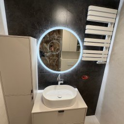 VASYL PAVLIUK Royal Remont - Nowoczesna łazienka z okrągłym lustrem LED, białą umywalką nablatową i grzejnikiem drabinkowym na tle ciemnej, marmurkowej ściany. Minimalistyczny design, jasne meble.