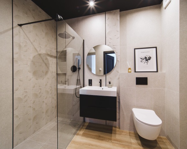 Beżowa łazienka z kabiną walk-in, okrągłym lustrem i drewnopodobną podłogą. Na ścianach dekoracyjne płytki z motywem kół. Czarna armatura i rama kabiny.