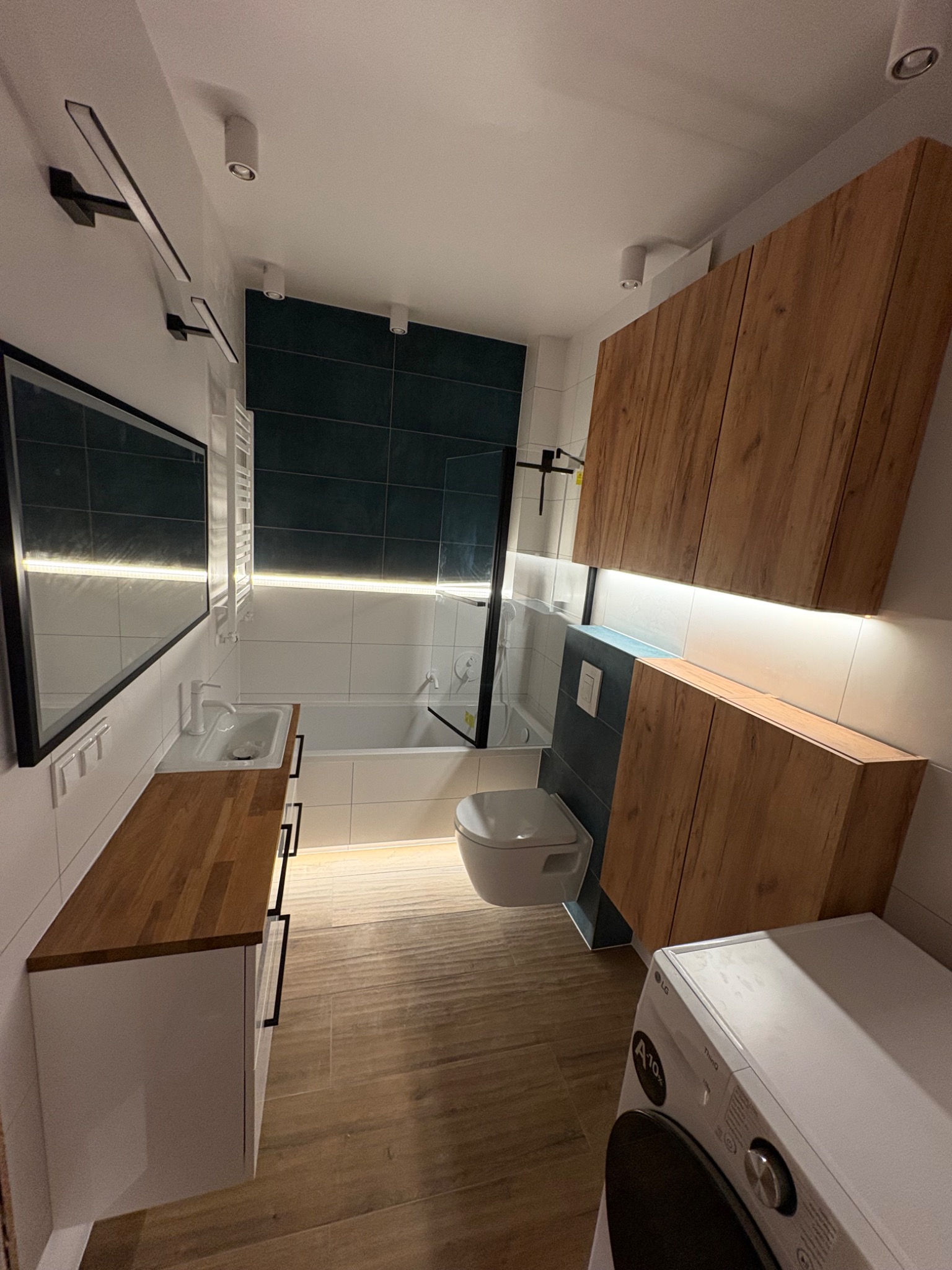 Nowoczesna łazienka z drewnianymi akcentami: szafki, blat umywalki. Uwagę przyciąga podświetlenie LED oraz ciemnozielona ściana nad wanną.