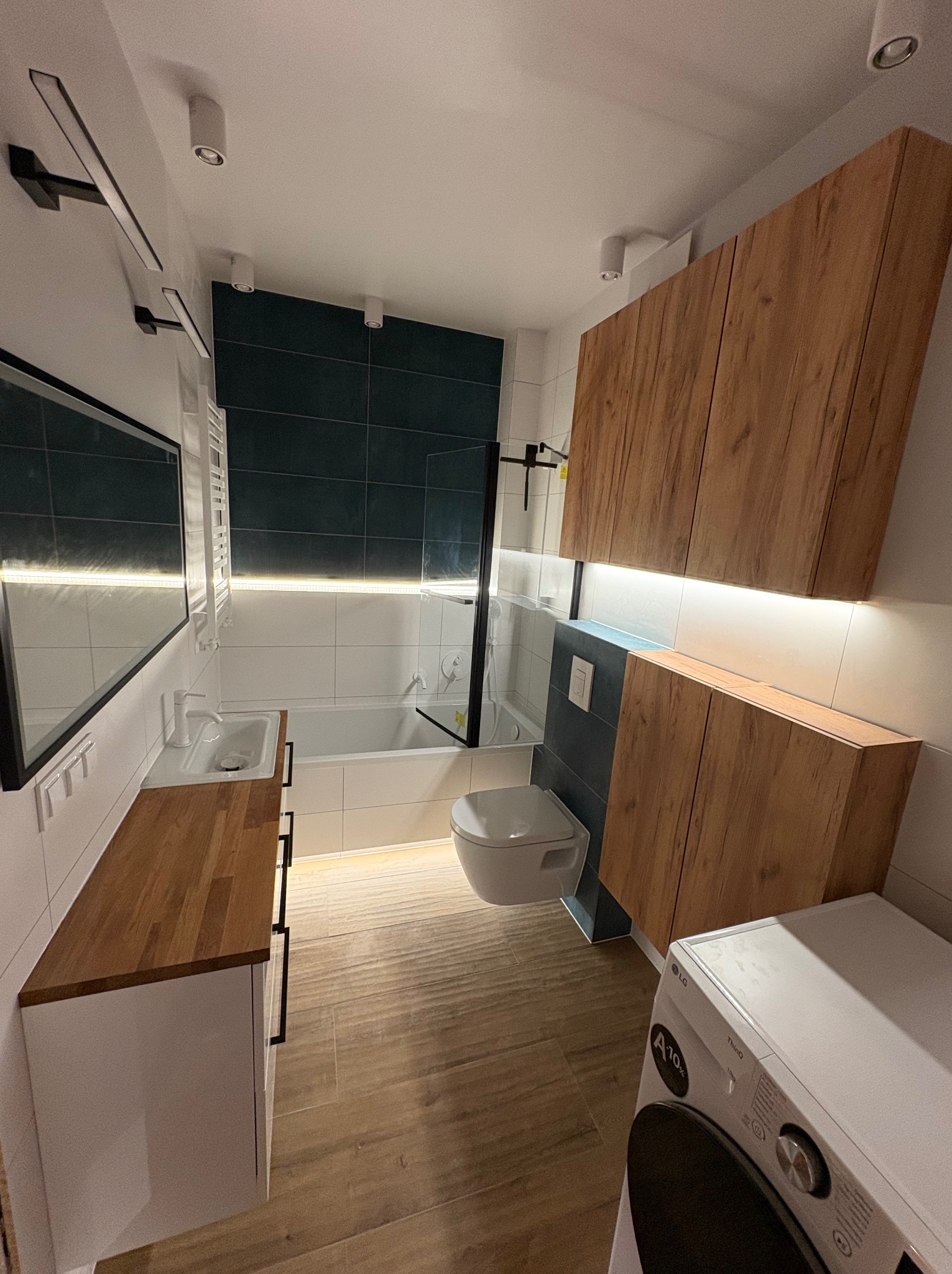 Nowoczesna łazienka po metamorfozie: drewniane szafki, minimalistyczna umywalka z blatem, ciemne płytki i subtelne oświetlenie LED tworzą spójny, elegancki design.