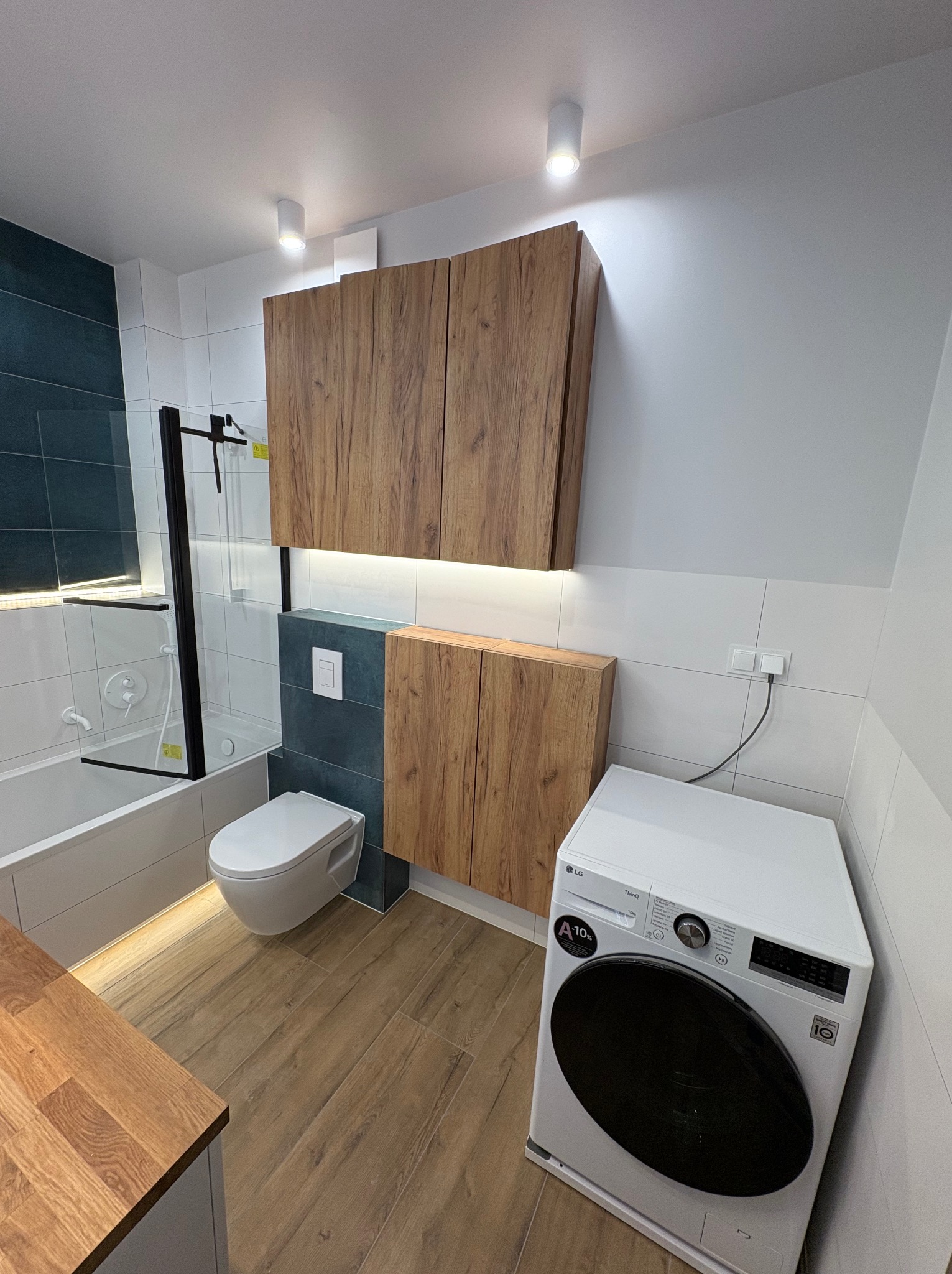 Nowoczesna łazienka z drewnianymi szafkami, kabiną prysznicową ze szkła, toaletą i pralką LG. Podświetlenie LED dodaje ciepła. Widok z perspektywy.